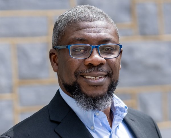 Adebayo "Bayo" Ogundipe in dark jacket and shirt without a tie, against a brick background