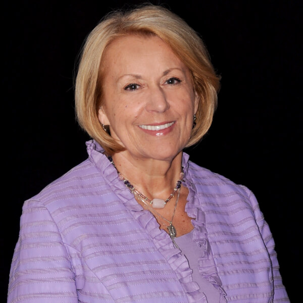 Kathy Crosby poses for a portrait. She is in a purple shirt against a black background.