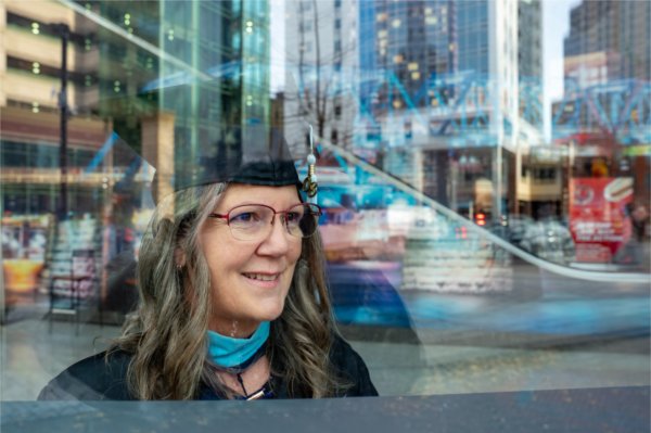  A person in academic regalia is seen through a window where a city scene is reflected behind them. 