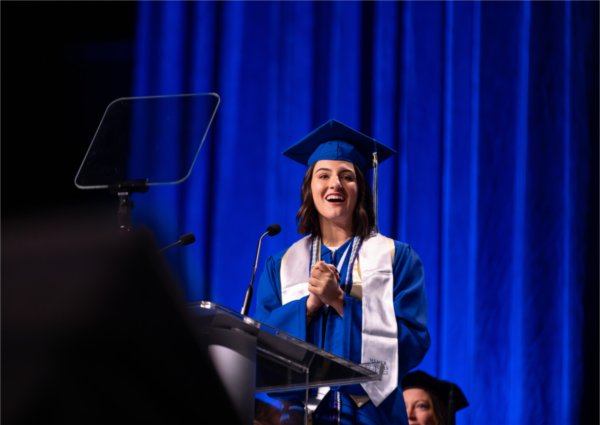  A student speaker wearing blue cap and gown speaks at the lectern. 