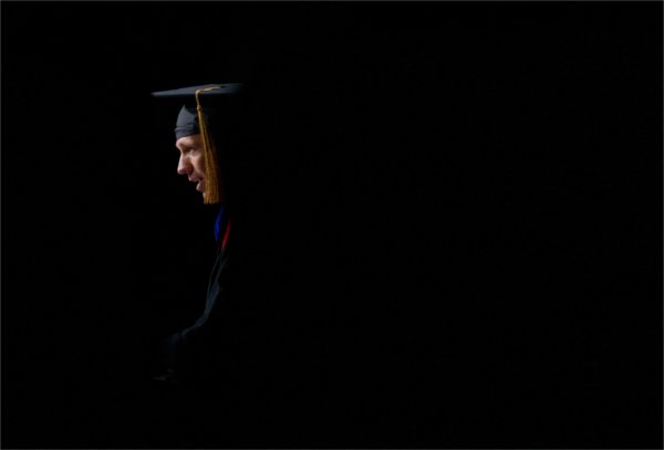  A speaker wearing a cap and gown with a golden tassel is highlighted by a beam of light.