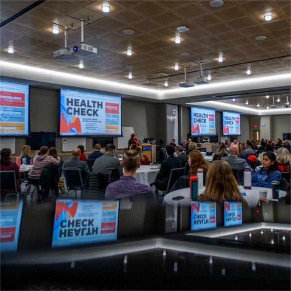 room of people seated at round tables, screens with Health Check around the room and reflected at bottom