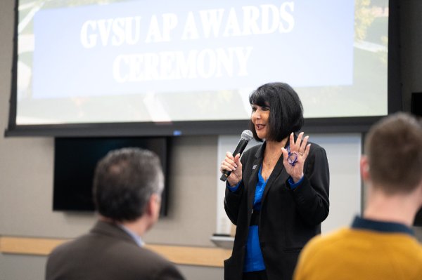 President Mantella speaks to the audience; a screen behind her reads GVSU AP Awards Ceremony