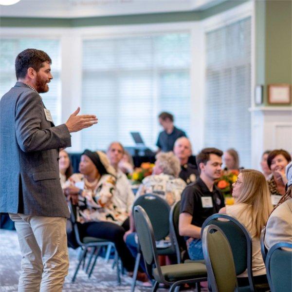 man stands and addresses room of people seated at round tables in Alumni House