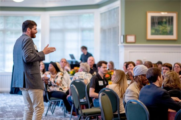 man stands to address audience seated at round tables in the Alumni House