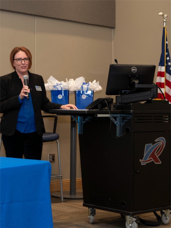 Kara Van Dam with microphone at front of room, hand resting on computer stand with blue gift bags behind her