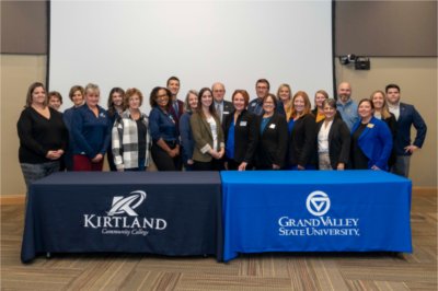 large group of people standing in two rows behind 2 tables draped with banners for GVSU and Kirtland Community College