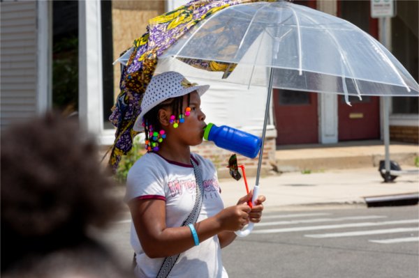A girl with colorful beads in her hair holds a water bottle in her mouth and holds an umbrella above her to shield herself from the sun.