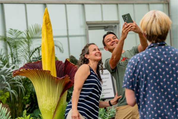Two people smile while taking a selfie. The corpse flower is in the background.