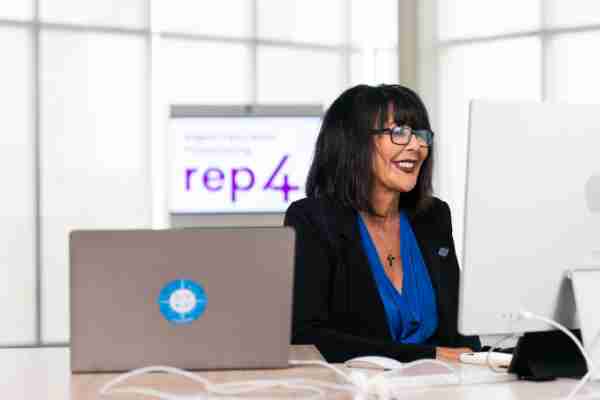 President Philomena V. Mantella smiles while looking at a computer screen. A screen with the REP4 logo is behind Mantella.